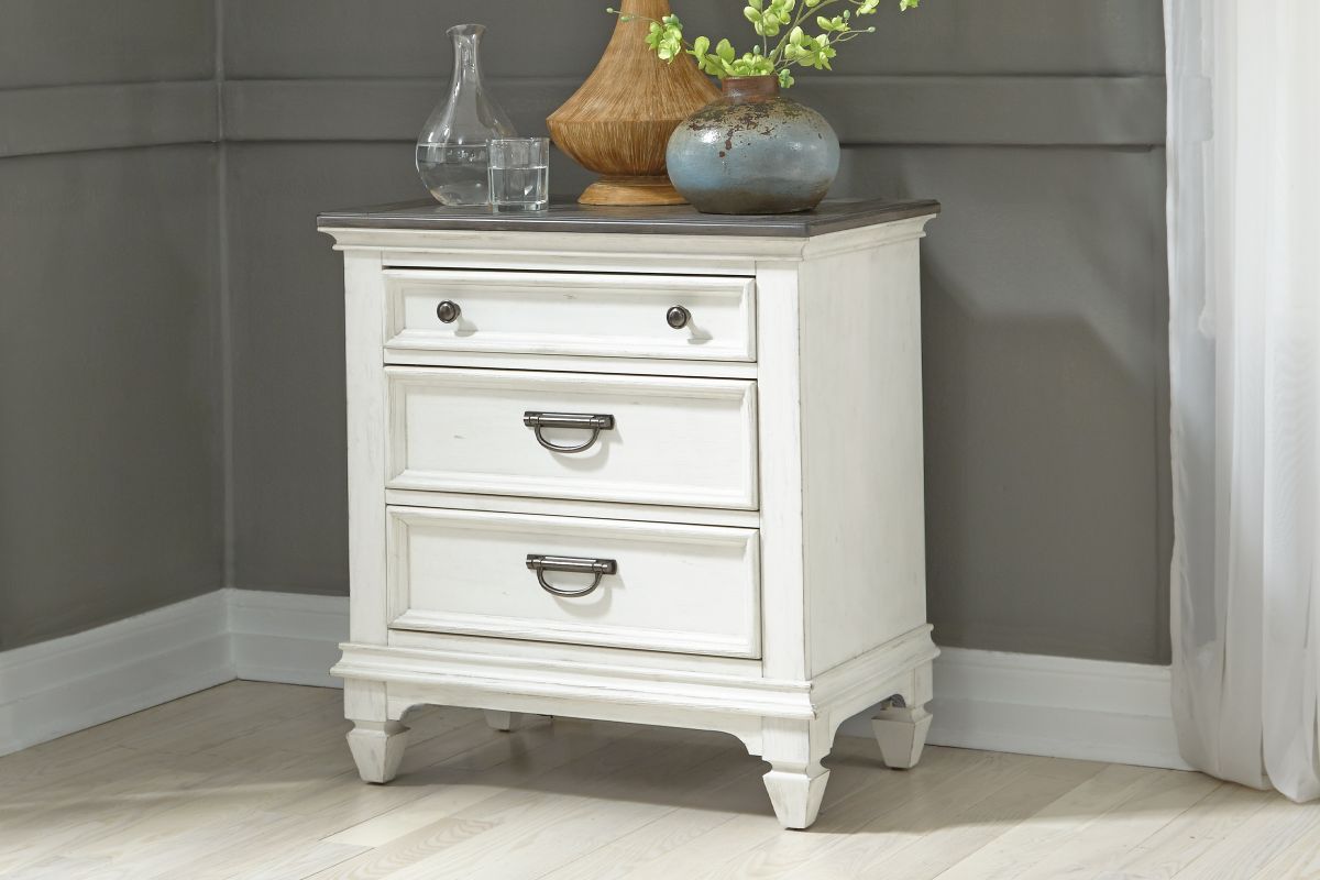 Corner view of a white wood 3-drawer nightstand against a gray wall